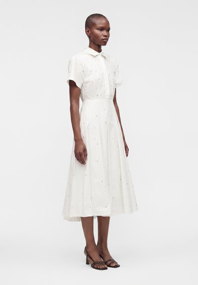 Femme portant une robe blanche à manches courtes, longueur mi-mollet, avec de petits détails brodés et des sandales noires à bouts ouverts, debout devant un fond uni.
