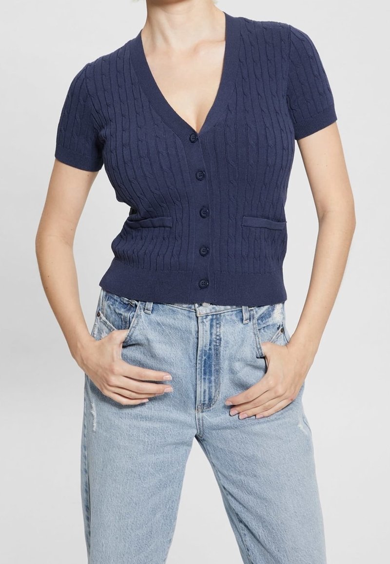 Femme portant un cardigan en maille torsadée bleu marine à manches courtes et boutonné, ainsi qu'un jean en denim bleu clair taille haute, les mains dans les poches.