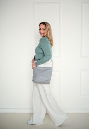 Gray shoulder bag with a smooth texture, featuring a zip closure and a detachable strap. The bag sits beside a person in light-colored pants.