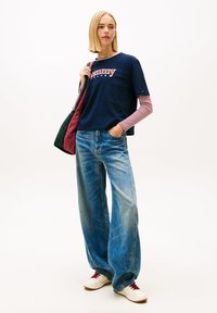 Granatowa koszula z krótkim rękawem z czerwonym logo „Tommy”, noszona na paski na długie rękawy, w połączeniu z szerokimi, wyblakłymi niebieskimi dżinsami i białymi trampkami.