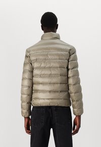 Beige dunjakke med en blank tekstur, horisontal quilting, høy krage og ribbede mansjetter. Kombinert med svarte jeans.