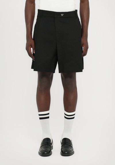 Schwarze gefaltete Shorts mit flachem Vorderteil, kombiniert mit weißen Socken mit schwarzen Streifen und schwarzen Slippern.