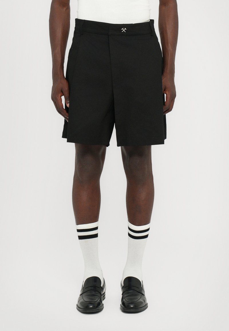Schwarze gefaltete Shorts mit flachem Vorderteil, kombiniert mit weißen Socken mit schwarzen Streifen und schwarzen Slippern.