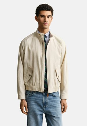 Man draagt een lichtbeige jas met rits over een blauw overhemd en gestreepte stropdas, gecombineerd met lichtblauwe jeans, staand tegen een effen achtergrond.