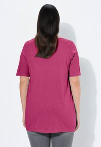 Różowa fioletowa koszulka z krótkim rękawem, okrągłym dekoltem i bocznymi rozcięciami, wykonana z gładkiego, lekkiego materiału. Widoczne spodnie w odcieniach szarości.