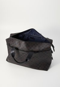Brown checkered duffel bag made of textured synthetic material. Features black handles and a spacious interior with a navy lining. Zipper closure.