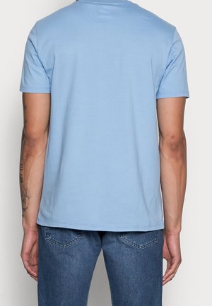 Homme de dos portant un t-shirt à manches courtes bleu clair uni et un jean en denim bleu, avec des tatouages visibles sur son bras gauche.