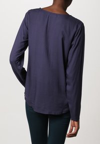 Långärmad blus i djup marinblå färg med en slät textur, rund halsringning och subtilt mönster. Sömmen är något längre bak.