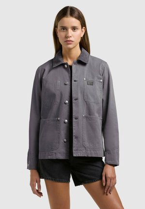 Jonge vrouw die een grijze jack met knopen en vier zakken aan de voorkant draagt, gecombineerd met een zwarte korte broek, kijkt recht vooruit met een neutrale gezichtsuitdrukking.