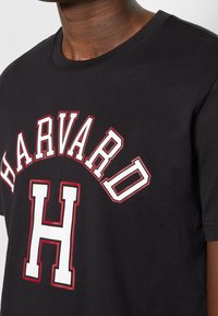 Close-up of person wearing black T-shirt with "HARVARD" in white and red outlined letters arched above a large block "H" on chest.