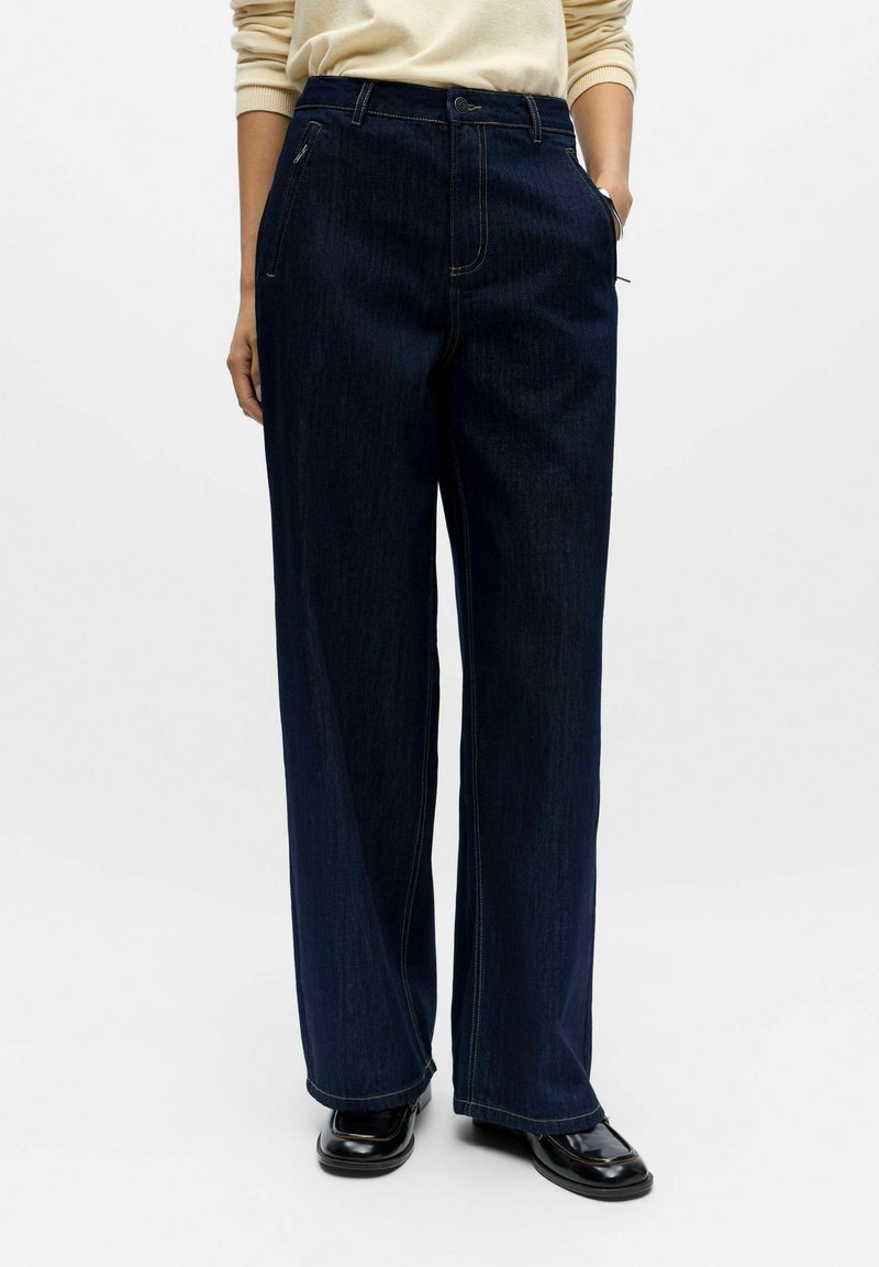 Tmavě modré džínsy s širokými nohavicemi z denimu, s vysokým pasem, předními kapsami a kontrastními švy. Hladká textura s čistým designem.