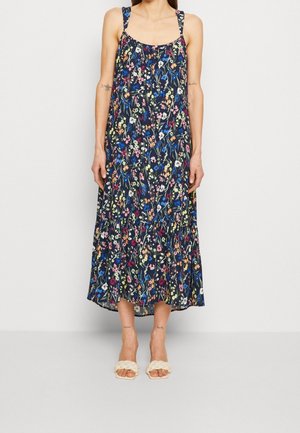 Mujer con un vestido negro sin mangas hasta los tobillos con estampado floral colorido y sandalias blancas trenzadas tipo slide.