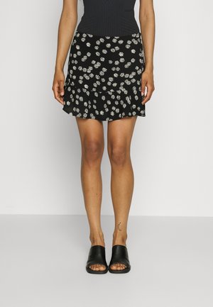 Mujer con falda negra con flores y margaritas blancas, top negro, sandalias negras abiertas y un pequeño tatuaje de luna creciente en la pierna.