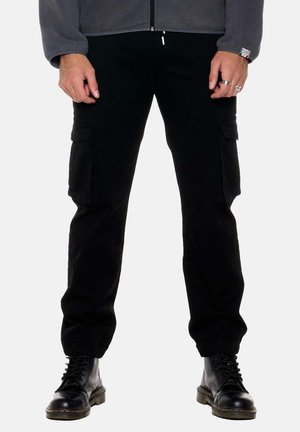 Homme portant un pantalon cargo noir avec des poches à rabat, des bottes noires à lacets et une veste grise, debout sur un fond blanc.