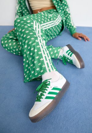 Personne portant un pantalon Adidas vert à motifs et des baskets Adidas Samba blanches avec des bandes vertes, assise sur un tapis bleu.