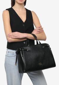 Sort læder tote taske med to håndtag. Har en struktureret overflade, cirkulære hardware detaljer og et logo på forsiden.