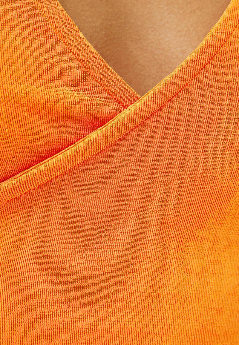 Primer plano de una persona que lleva una prenda naranja texturizada con escote en V y un detalle de tela superpuesta en el pecho.