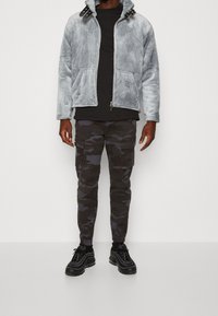 Szara kurtka z imitacji futra zapinana na zamek, z czarnymi akcentami, w połączeniu z ciemnymi spodniami cargo w kamuflażu oraz czarnymi butami sportowymi.