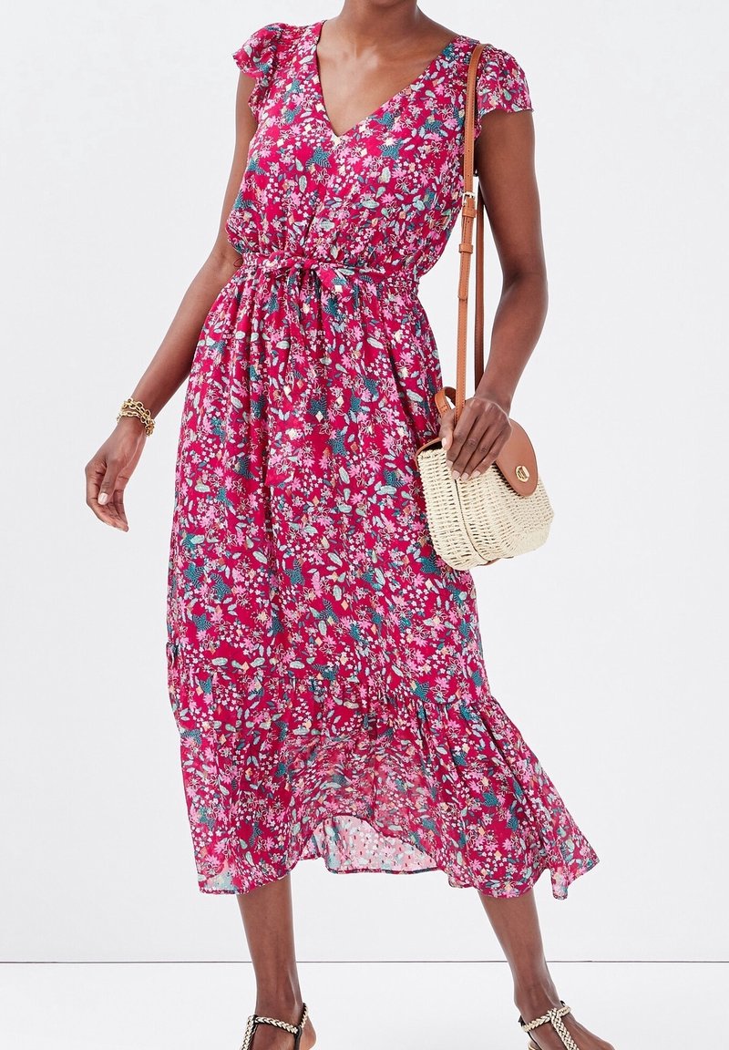 Robe longue fleurie rose, avec des manches courtes à volants, une taille cintrée et un ourlet à volants. Associée à un petit sac bandoulière tressé.