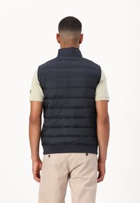 Navy puffer vest featuring a high collar, horizontal quilted design, elastic hem, and worn over a light-colored short-sleeve shirt.