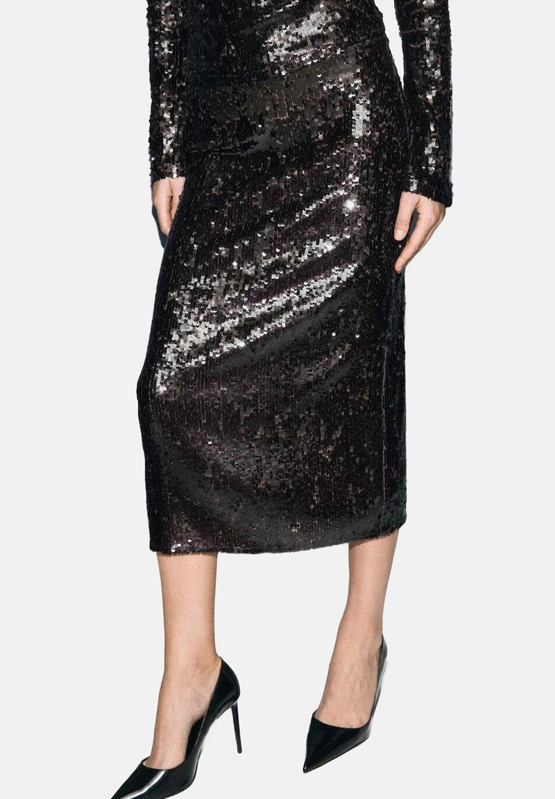 Robe noire en sequins avec des manches longues et une silhouette cintrée, descendant jusqu'au mollet. Portée avec des escarpins noirs à bout pointu.