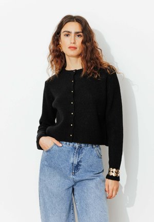 Cardigan noir côtelé avec des boutons dorés, col rond et manches longues. Accent doré remarquable au poignet. Porté sur un jean taille haute bleu clair.