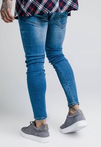 Persona que lleva jeans skinny azules, zapatillas de ante grises, una camisa de cuadros y muestra tatuajes en su mano izquierda y tobillo derecho.