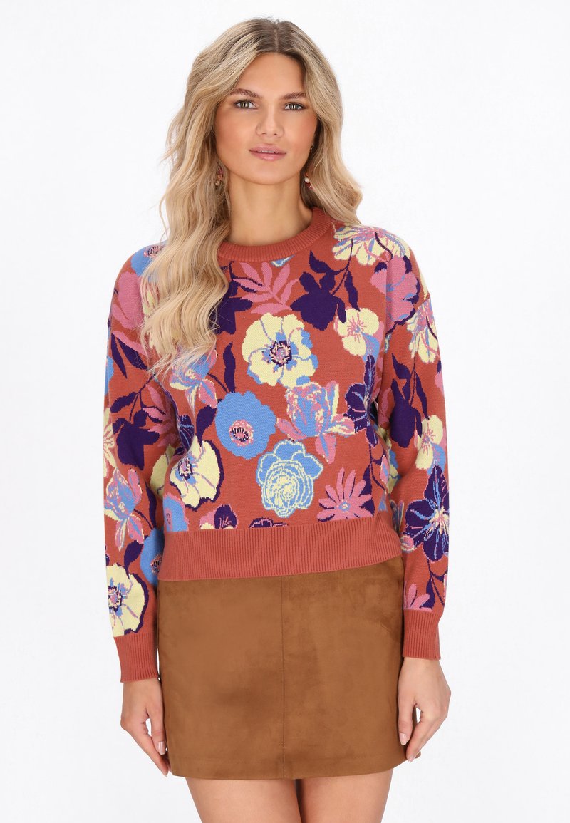 Maglione lavorato a maglia con motivi floreali multicolore su uno sfondo ruggine, con maniche lunghe e scollatura a girocollo a costine. Abbinato a una gonna marrone.