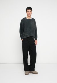 Νέος άνδρας στέκεται, φοράει σκούρο γκρι χνουδωτό πουλόβερ, μαύρο πλισέ παντελόνι και μπεζ σουέτ μπότες, με φόντο απλό λευκό τοίχο.