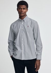 Randig skjorta med knäppning framtill, tunna marinblå och vita ränder, knäppkrage och en liten broderad logotyp på bröstet.