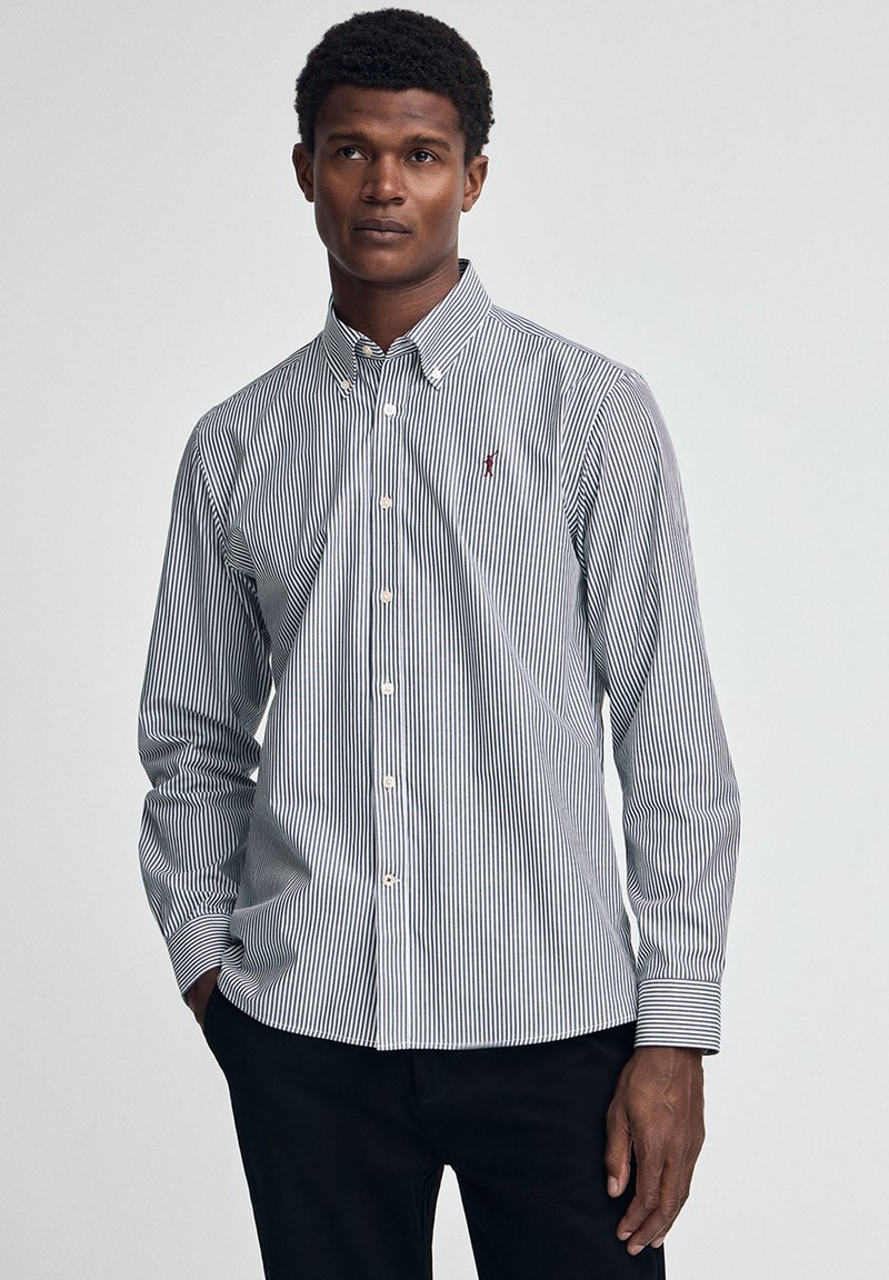 Randig skjorta med knäppning framtill, tunna marinblå och vita ränder, knäppkrage och en liten broderad logotyp på bröstet.