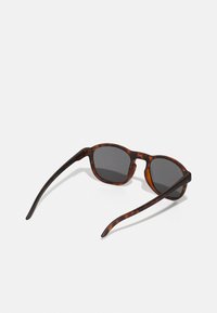 Óculos de sol em padrão tartaruga com lentes pretas. Design de armação arredondada com um acabamento suave. Templos finos que se estendem para fora.
