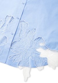 Lichtblauw shirt met witte bloemenborduring langs de zoom en het revers, met knoopsluiting en een gestructureerde stoftextuur.