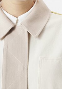 Manteau beige et crème avec un grand col, des détails de couture diagonale et une poche avant avec des coutures renforcées. Texture lisse.