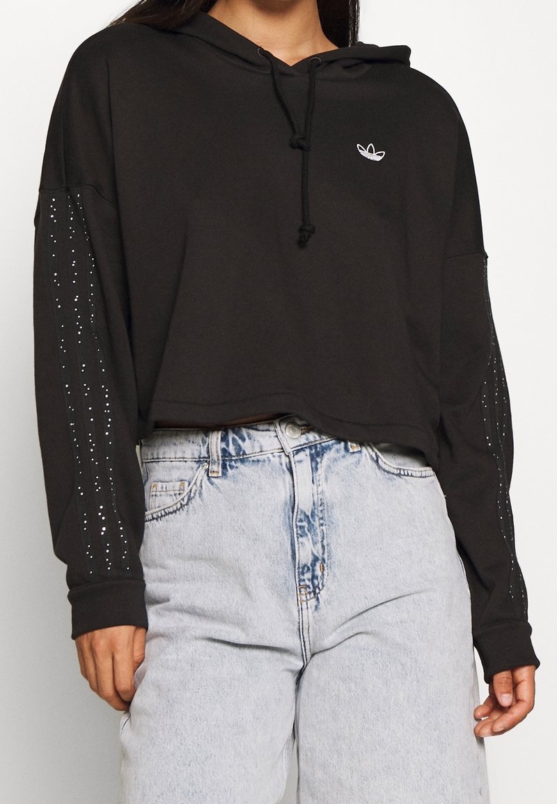 Felpa nera corta con cappuccio e coulisse, caratterizzata da maniche lunghe decorate con strass e un piccolo logo sul petto. Abbinata a jeans chiari.