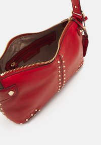 Czerwona skórzana torebka z ozdobami z złotych ćwieków, zamkiem błyskawicznym na górze i wewnętrzną kieszenią. Posiada odpinany pasek na ramię.