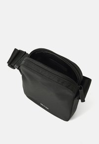 Borsa a tracolla nera in tessuto con zip sulla parte superiore, forma rettangolare piatta e tracolla rimovibile. Presenta un logo sul fronte e una texture liscia.