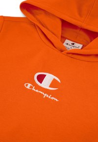 Sudadera de algodón naranja con un bolsillo frontal. Logo de Champion bordado en blanco y rojo. Textura suave con un ajuste holgado.