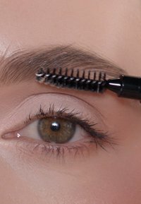 Ein schwarzer Augenbrauenpinsel kämmt durch braune Augenbrauenhaare; das Auge hat lange Wimpern und ein leichtes, glattes Augenlid. Die Haut sieht makellos aus.