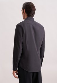 Camicia nera in tessuto intrecciato con maniche lunghe, colletto e polsini con bottoni. La parte posteriore presenta una piega centrale e un orlo curvo. Texture liscia.