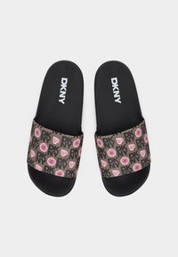 Sandales à glissière noires avec des lanières à motif marron ornées de cœurs roses, de fleurs et de logos DKNY, texte DKNY blanc sur les semelles.