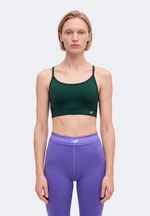 Une femme présente un soutien-gorge de sport vert foncé et un legging violet sur un fond uni et clair.