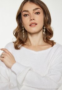 Biały, textured sweter z dekoltem w kształcie V i długimi, luźnymi rękawami. Na zdjęciu znajduje się ręka nosząca srebrny pierścionek oraz zbliżenie na złote i perłowe kolczyki.