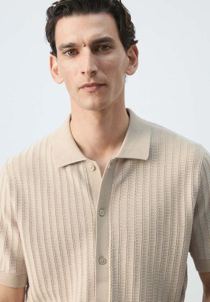 Homme aux cheveux foncés portant un polo en maille beige à manches courtes avec un motif vertical texturé, regardant droit devant.