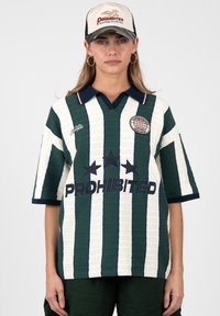 Mujer que lleva una camiseta de rayas verdes y blancas con el texto "PROHIBITED" y una gorra beige con el mismo logo, de pie frente a un fondo liso.