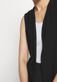 Svart ärmlös väst med öppen front, tillverkad av ett slätt tyg. Buren över en vit ribbad tank top med avslappnad passform.