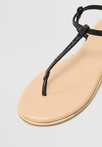 Juodas thong sandal su metalizuotu auksiniu padu, turintis tekstūruotą juodą dirželį ir glotnų šviesiai bežinį pado paviršių.