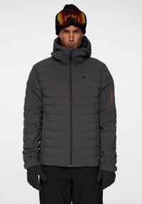 Grå vadderad jacka med hög krage, framdragkedja och röd logotyp på ärmen. Bärs med en svart mössa och färgglada skidglasögon.