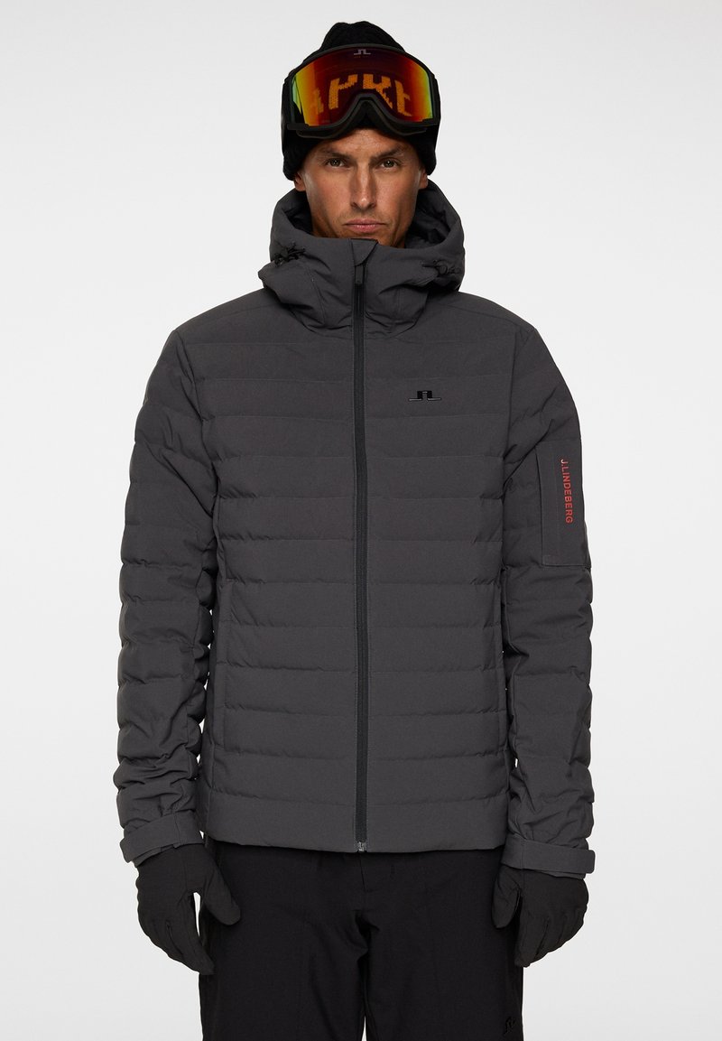 Grå vadderad jacka med hög krage, framdragkedja och röd logotyp på ärmen. Bärs med en svart mössa och färgglada skidglasögon.