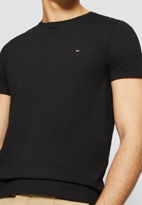 T-shirt en coton noir avec un col rond, des manches courtes et un petit logo brodé en rouge, blanc et bleu sur la poitrine gauche.